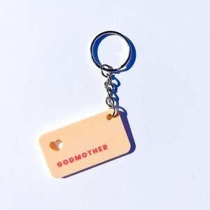 May include: A peach-colored keychain with a heart cut-out and the word "GODMOTHER" in pink lettering.