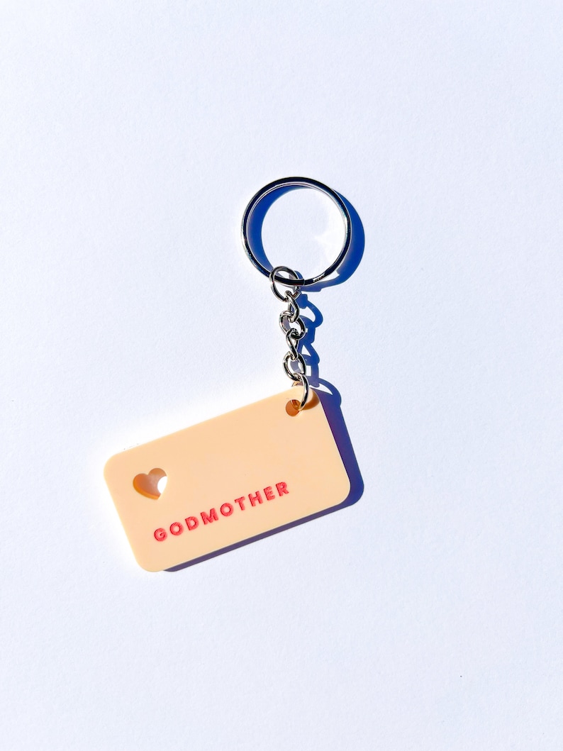 May include: A peach-colored keychain with a heart cut-out and the word "GODMOTHER" in pink lettering.