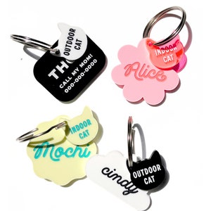 May include: Four cat ID tags with different names and colors.  The tags are shaped like a square, a flower, a cloud, and a paw print.  The tags have black, white, pink, and yellow backgrounds.  The text on the tags reads "Alice", "Mochi", "Cindy", and "Thor".  The tags also have small cat silhouettes with the words "Indoor Cat" and "Outdoor Cat" on them.