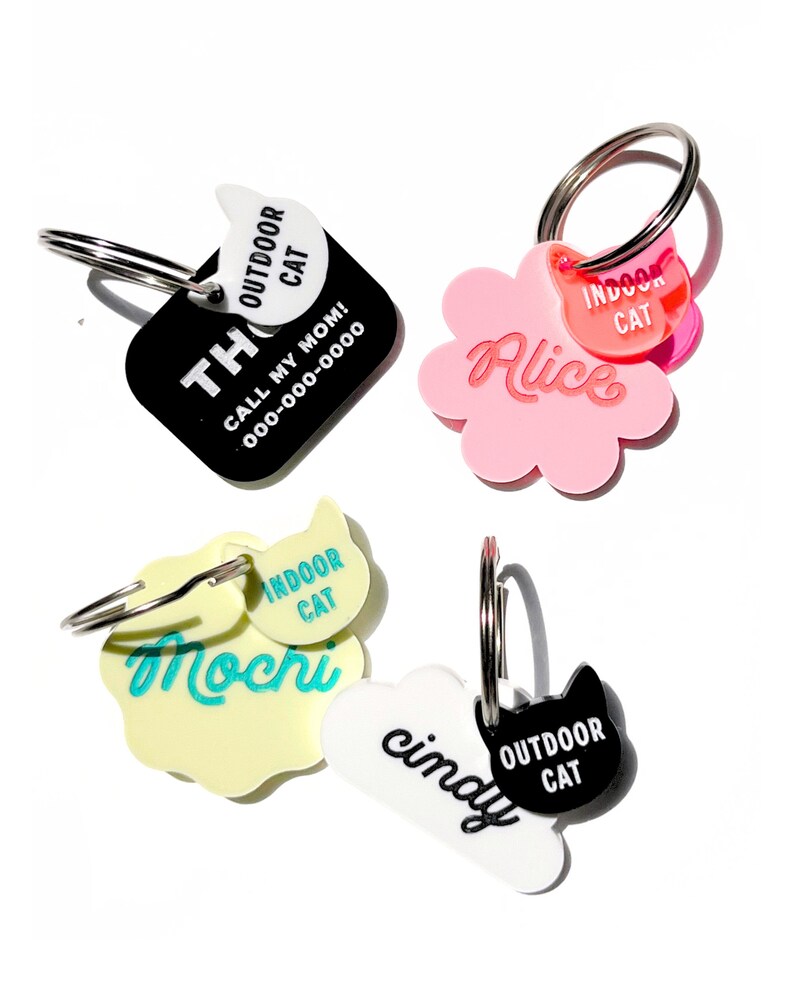 May include: Four cat ID tags with different names and colors.  The tags are shaped like a square, a flower, a cloud, and a paw print.  The tags have black, white, pink, and yellow backgrounds.  The text on the tags reads "Alice", "Mochi", "Cindy", and "Thor".  The tags also have small cat silhouettes with the words "Indoor Cat" and "Outdoor Cat" on them.