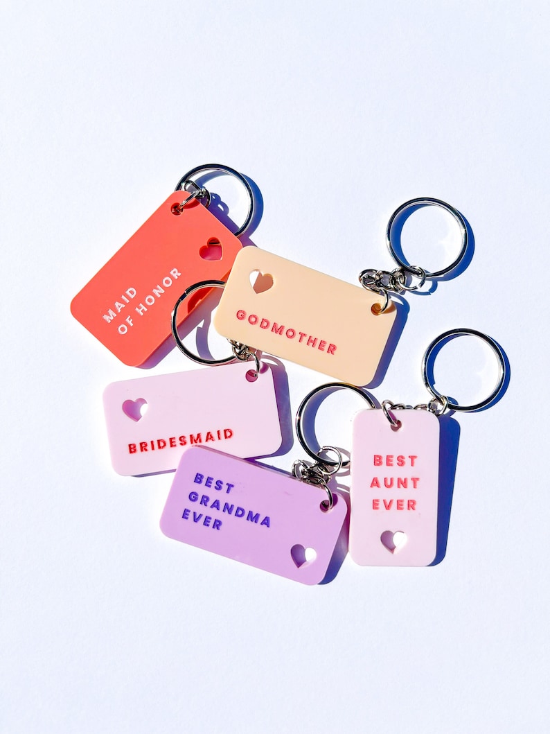 May include: Five colorful keychains with different titles, including "Maid of Honor", "Godmother", "Bridesmaid", "Best Grandma Ever", and "Best Aunt Ever". Each keychain has a heart-shaped cutout and a silver key ring.