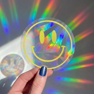 Peut inclure: Un autocollant circulaire présentant le contour d'un visage souriant jaune sur un fond holographique qui reflète des motifs lumineux arc-en-ciel. Le motif se compose de deux yeux ovales et d'une bouche incurvée, le tout en trait jaune.