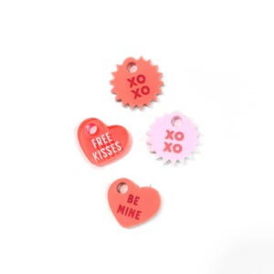 Op de afbeelding: Vier Valentijnsdag-thema acrylcharms in de vorm van harten en een ster. De charms zijn roze en rood en hebben de volgende tekst: "Free Kisses", "Be Mine" en "XO" op twee van de charms.