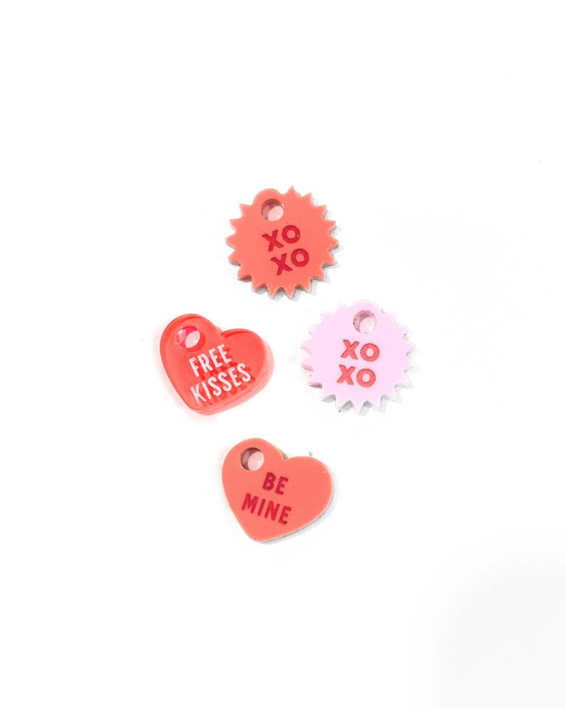 Op de afbeelding: Vier Valentijnsdag-thema acrylcharms in de vorm van harten en een ster. De charms zijn roze en rood en hebben de volgende tekst: "Free Kisses", "Be Mine" en "XO" op twee van de charms.