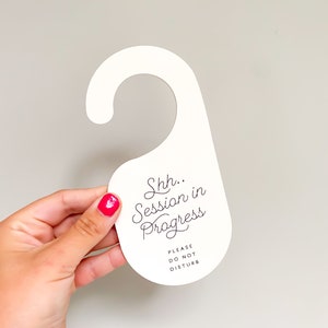 May include: A white door hanger with a hook-shaped top. The text "Shh.. Session in Progress" is printed in a black cursive script, followed by the text "PLEASE DO NOT DISTURB" in a smaller, simple black sans-serif font centred below it.