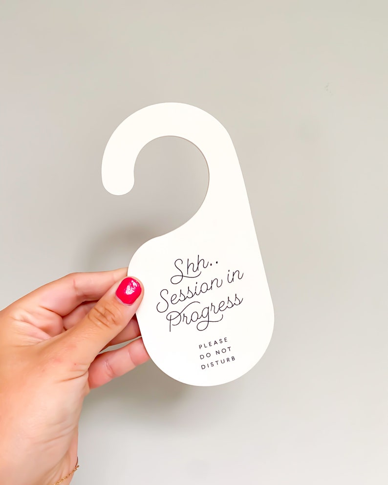 May include: A white door hanger with a hook-shaped top. The text "Shh.. Session in Progress" is printed in a black cursive script, followed by the text "PLEASE DO NOT DISTURB" in a smaller, simple black sans-serif font centred below it.