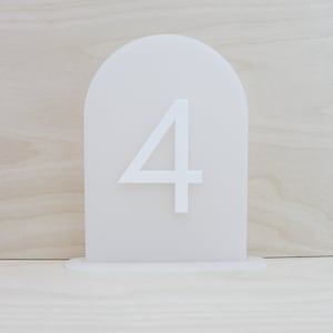 May include: White acrylic table number with the number 4 printed on it. The number is on a stand with a rounded top.