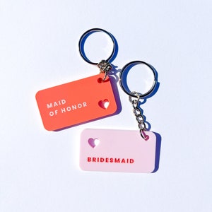 May include: Two keychains with pink and orange rectangular tags. The orange tag says "Maid of Honor" with a heart cutout. The pink tag says "Bridesmaid" with a heart cutout.