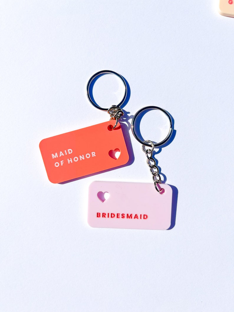 May include: Two keychains with pink and orange rectangular tags. The orange tag says "Maid of Honor" with a heart cutout. The pink tag says "Bridesmaid" with a heart cutout.