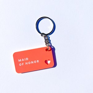 May include: A bright orange keychain with a white heart cutout and the text "Maid of Honor" in white lettering.