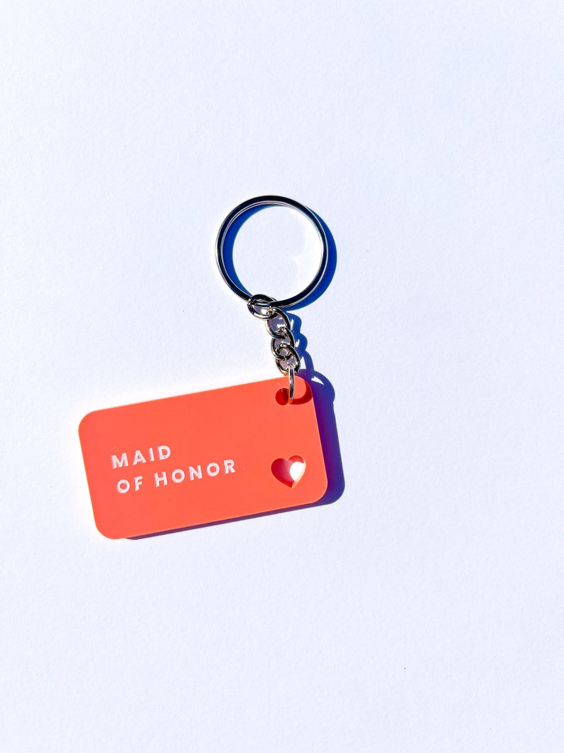 May include: A bright orange keychain with a white heart cutout and the text "Maid of Honor" in white lettering.