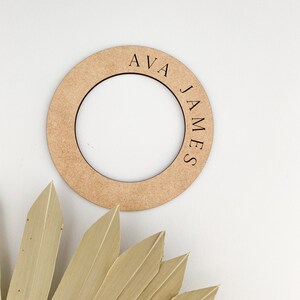 May include: A round, light brown wooden sign with the names "AVA" and "JAMES" engraved on it. The sign has a circular cutout in the center. A dried, beige palm leaf is in the lower left corner, against a white background.