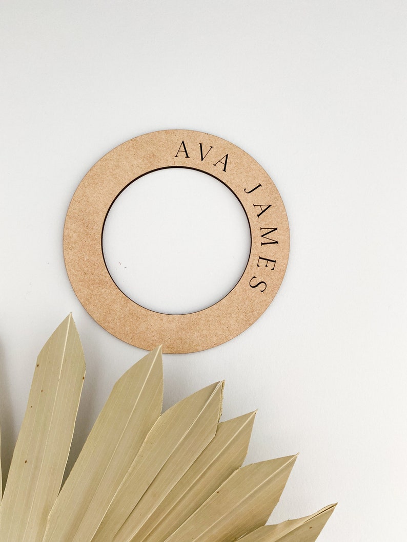 May include: A round, light brown wooden sign with the names "AVA" and "JAMES" engraved on it. The sign has a circular cutout in the center. A dried, beige palm leaf is in the lower left corner, against a white background.