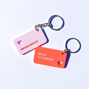 May include: Two keychains with heart cutouts. One is pink with the text "BRIDESMAID" and the other is orange with the text "MAID OF HONOR".