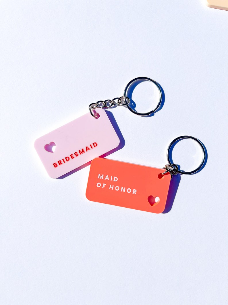 May include: Two keychains with heart cutouts. One is pink with the text "BRIDESMAID" and the other is orange with the text "MAID OF HONOR".