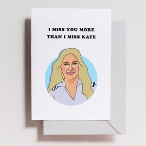 May include: A white card with black text that reads "I miss you more than I miss Kate". A cartoon illustration of a woman with blonde hair wearing a white shirt with black stripes on the shoulders is in the center of the card.