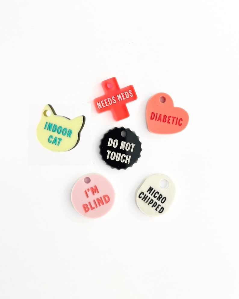 May include: Six colorful plastic pet ID tags with different messages. The tags are shaped like a cat, a heart, a cross, a circle, and an oval. The messages on the tags are "Indoor Cat", "Diabetic", "Needs Meds", "Do Not Touch", "I'm Blind", and "Microchipped".