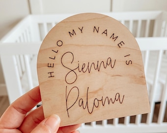 May include: An arch-shaped light wood plaque featuring the engraved text "HELLO MY NAME IS" in a sans-serif font, with the name "Sienna Paloma" written in a cursive script below. The plaque has a smooth, natural wood grain finish.