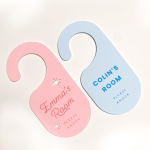 May include: Two door hanger signs, one pink and one pale blue. The pink sign reads "Emma's Room PLEASE KNOCK" with starburst accents. The blue sign reads "COLIN'S ROOM PLEASE KNOCK".