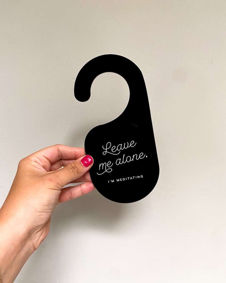 May include: A black door hanger with a hook shape at the top. The front features white text that reads Leave me alone. in a cursive script, followed by the text I'M MEDITATING in a simple sans-serif font centred below the cursive script.