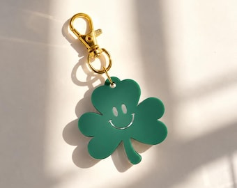 May include: A green shamrock-shaped keychain featuring a white engraved smiley face. The clover charm is attached to a gold-toned metal lobster clasp, providing a secure way to attach the accessory to bags, keys, or other personal items.