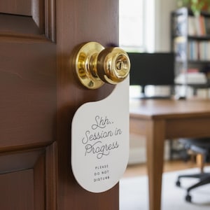 May include: A white door hanger with a curved top and a circular cutout, hanging from a brass doorknob on a dark wood door. The text reads Shh.. Session in Progress and below that, in smaller capital letters, it says PLEASE DO NOT DISTURB.