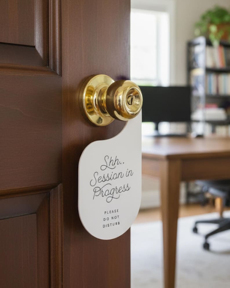 May include: A white door hanger with a curved top and a circular cutout, hanging from a brass doorknob on a dark wood door. The text reads Shh.. Session in Progress and below that, in smaller capital letters, it says PLEASE DO NOT DISTURB.