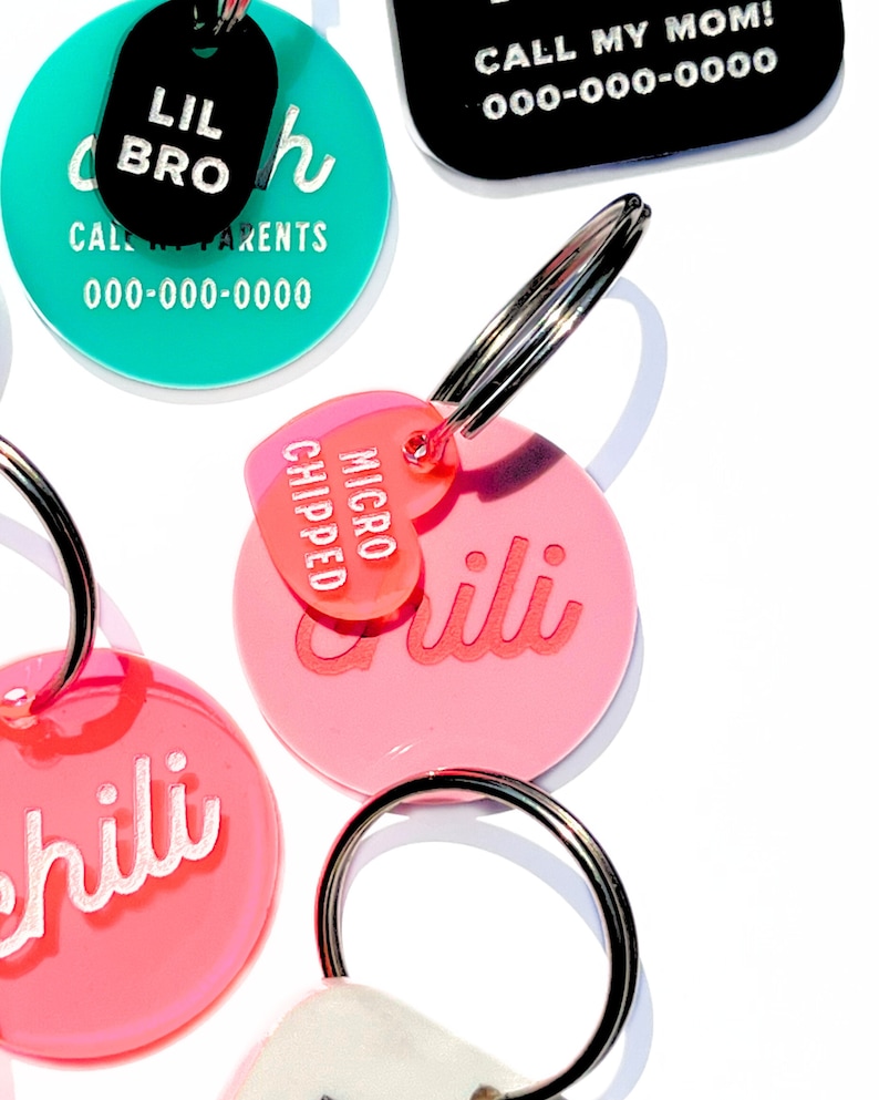 May include: Four colorful pet ID tags with key rings. The tags are round and have white text on a colored background. The tags read "Lil Bro", "Call Parents 000-000-0000", "Chili", and "Chili Micro Chipped".