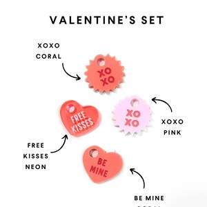 Op de afbeelding: Een set van vier acrylcharms met een Valentijnsdagthema. De charms zijn in de vorm van harten en sterren en bevatten de zinnen "XOXO", "Free Kisses", "Be Mine" en "XOXO". De charms zijn in de kleuren koraal, roze en neon.