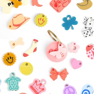 May include: A collection of colorful plastic charms with various designs, including a bone, a crown, a peace sign, a turtle, a dice, a heart, a butterfly, a sun, a sock, a bow, a peach, a seashell, a star, a tennis ball, a clover, a cat, a duck, a ladybug, a cowboy hat, a dice, and a flower.