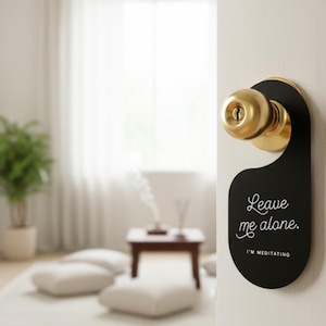 May include: A black door hanger with a rounded bottom and a circular cutout at the top, hanging from a brass-coloured doorknob. The text "Leave me alone." is printed in white script, with "I'M MEDITATING" in white block letters underneath.