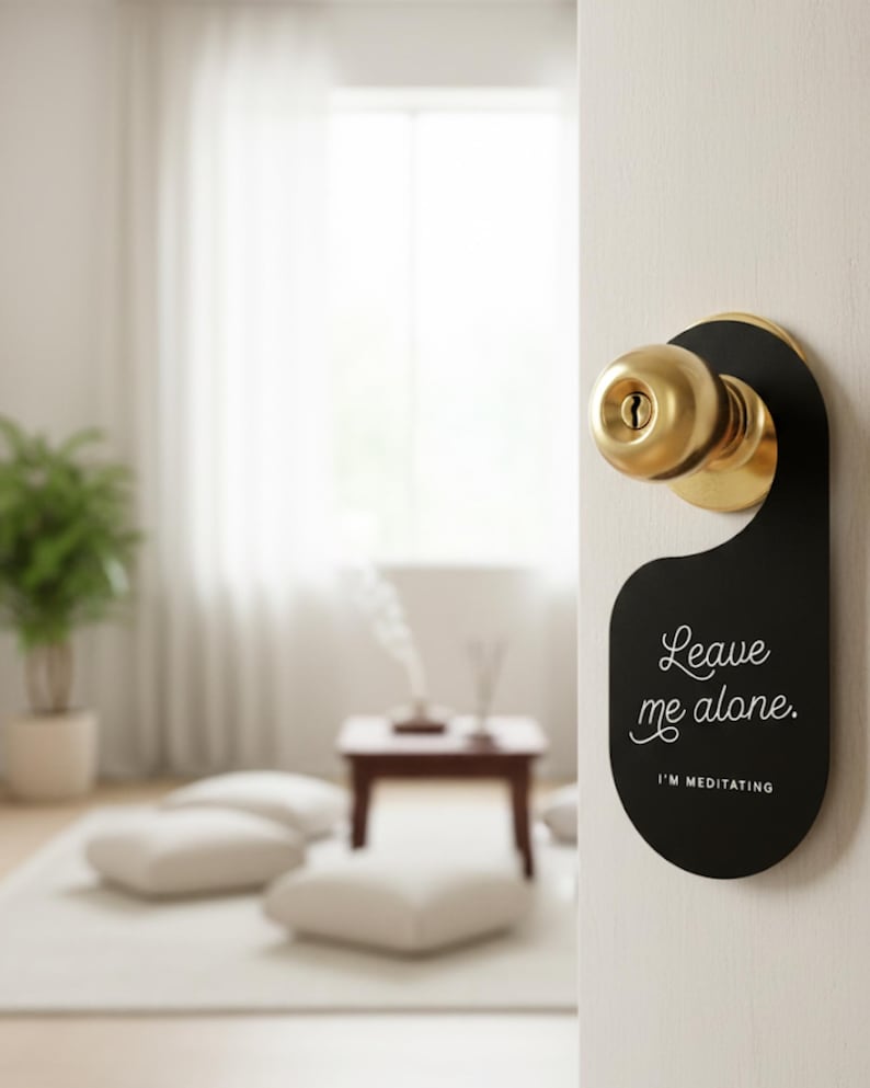 May include: A black door hanger with a rounded bottom and a circular cutout at the top, hanging from a brass-coloured doorknob. The text "Leave me alone." is printed in white script, with "I'M MEDITATING" in white block letters underneath.