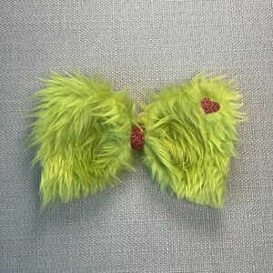 May include: A bright lime green faux fur bow accessory featuring a textured, shaggy appearance. The centre of the bow is cinched with a small, rectangular red glitter accent, and a matching red glitter heart is positioned on the right side.