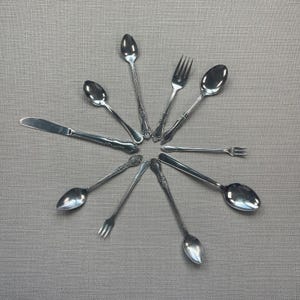 May include: A collection of nine silver-toned metal utensils arranged in a circular pattern, including various spoons, forks, and a single dinner knife. Each piece features a decorative embossed handle design with a polished, reflective finish.