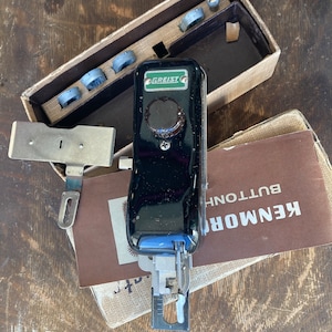 May include: A black and green buttonhole sewing machine with a knob and a metal foot. The machine is in its original box with a brown paper label that reads "KENMORB BUTTONH.