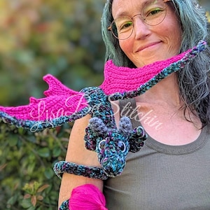 May include: A crocheted dragon toy featuring a variegated dark blue, green, and black yarn body with bright pink wings and a pink tail tuft. The dragon has small grey horns and black eyes, designed to wrap around an arm. Text reads Lisa Be Stitchin.