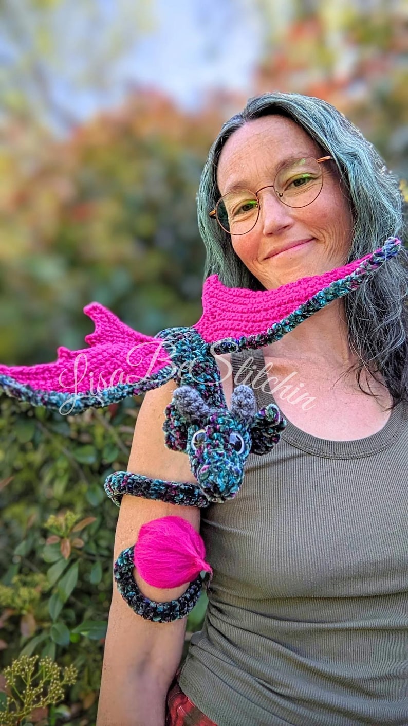 May include: A crocheted dragon toy featuring a variegated dark blue, green, and black yarn body with bright pink wings and a pink tail tuft. The dragon has small grey horns and black eyes, designed to wrap around an arm. Text reads Lisa Be Stitchin.