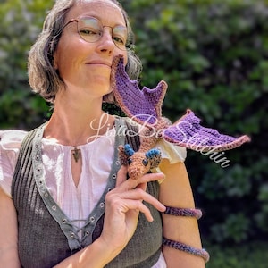 May include: A small, handcrafted crochet dragon toy featuring a tan body, blue ear accents, and large purple wings. The dragon has a long, coiled tail that wraps around an arm. The text Lisa B's Stitches is overlaid across the centre of the image.