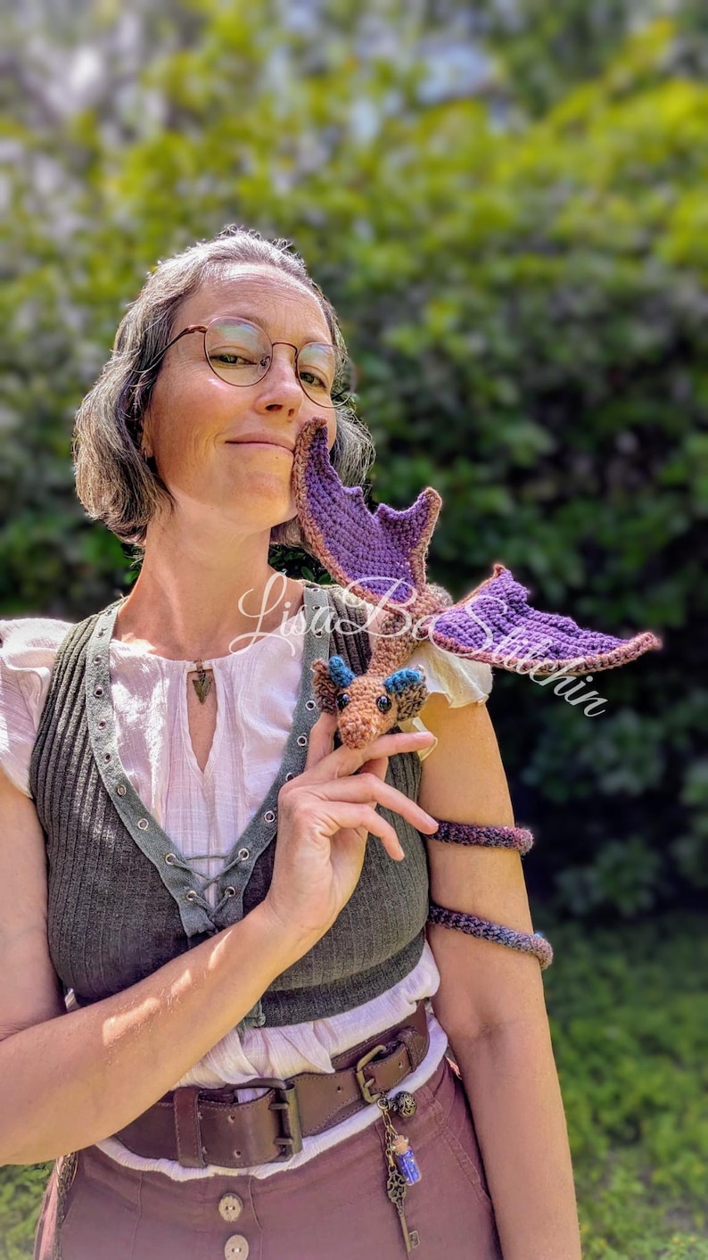 May include: A small, handcrafted crochet dragon toy featuring a tan body, blue ear accents, and large purple wings. The dragon has a long, coiled tail that wraps around an arm. The text Lisa B's Stitches is overlaid across the centre of the image.