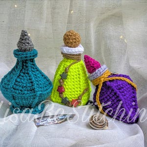 Puede incluir: Tres botellas de poción de ganchillo hechas a mano en verde azulado, verde lima y morado. La botella verde azulado tiene una tapa gris, la botella verde tiene una tapa marrón y detalles de estrellas, y la botella morada tiene un sombrero rosa y blanco. Las botellas están sobre una superficie blanca.
