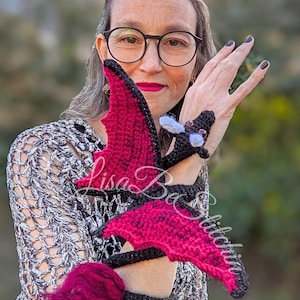 May include: A crocheted dragon toy featuring a black body with vibrant red wings and a matching red tufted tail. The dragon is designed to wrap around an arm, with white accents on the head and the text Lisa Be Stitched overlaid on the image.