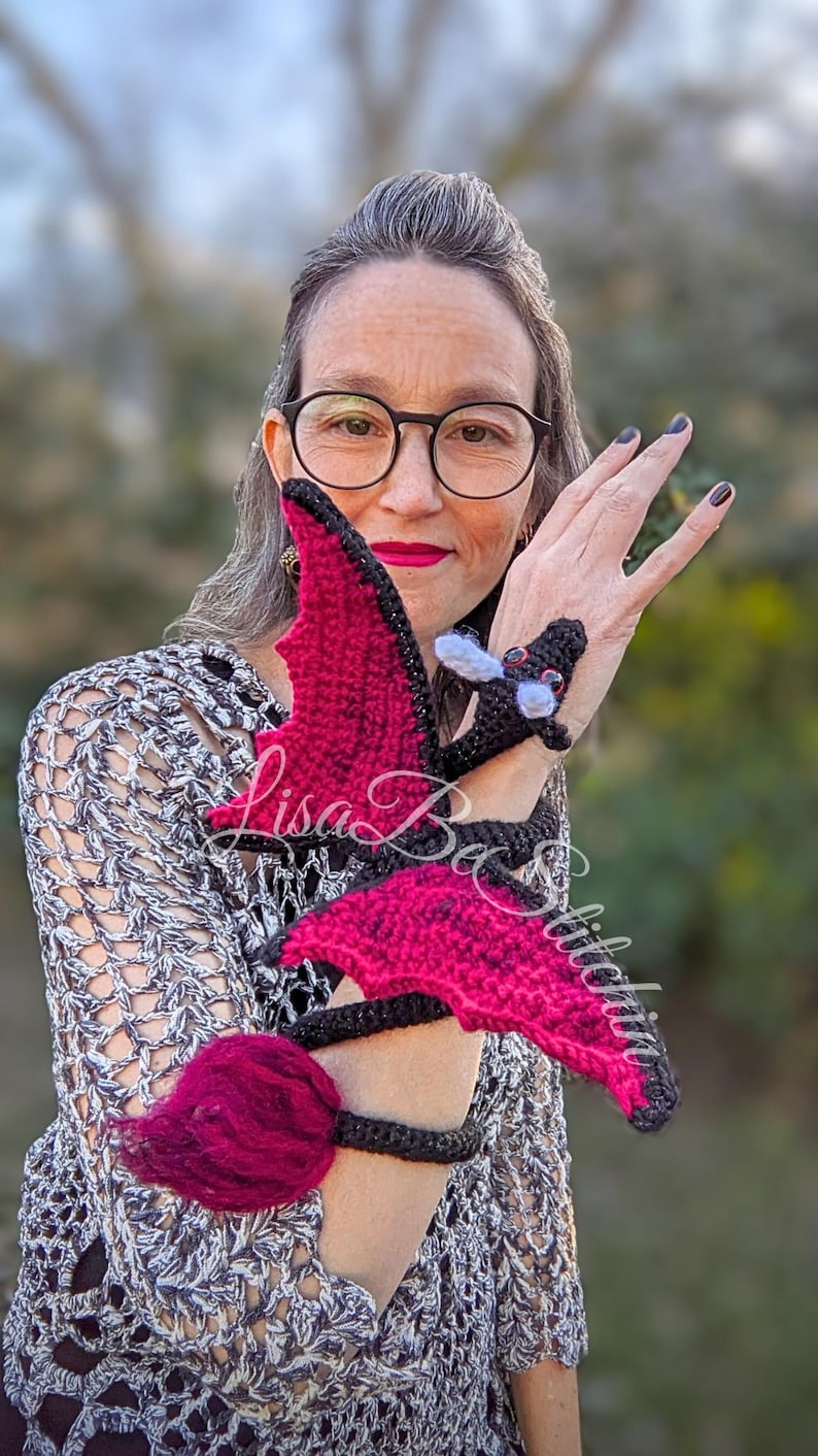May include: A crocheted dragon toy featuring a black body with vibrant red wings and a matching red tufted tail. The dragon is designed to wrap around an arm, with white accents on the head and the text Lisa Be Stitched overlaid on the image.