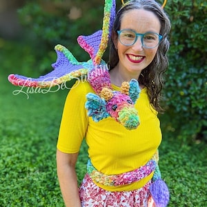 May include: A multi-coloured crochet dragon plush toy with a long body, wings, and a tail, featuring a vibrant mix of purple, green, yellow, and pink yarn. The dragon is designed to wrap around the shoulders and torso, with a textured, soft appearance.