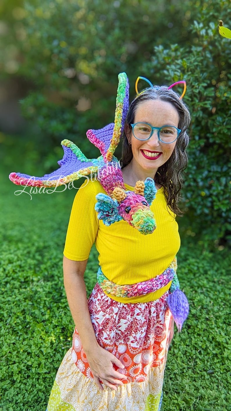 May include: A multi-coloured crochet dragon plush toy with a long body, wings, and a tail, featuring a vibrant mix of purple, green, yellow, and pink yarn. The dragon is designed to wrap around the shoulders and torso, with a textured, soft appearance.