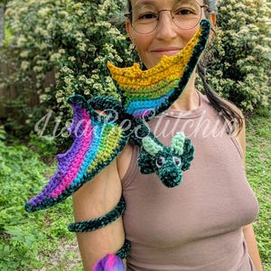 May include: A dark green crocheted dragon plush toy with rainbow-coloured wings and a long, curled tail ending in a vibrant pink, blue, and purple tuft. The dragon features small horns, black safety eyes, and a textured yarn body construction.