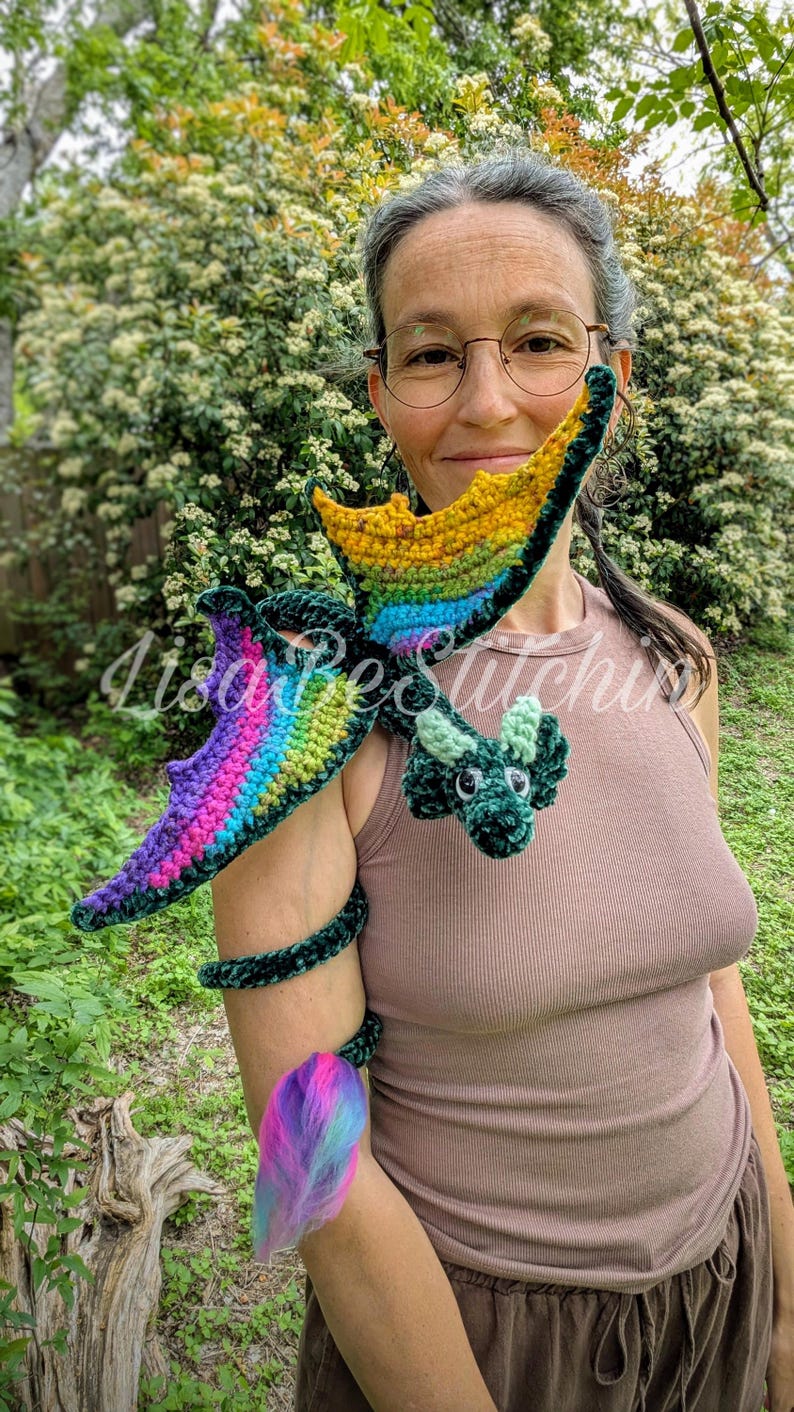 May include: A dark green crocheted dragon plush toy with rainbow-coloured wings and a long, curled tail ending in a vibrant pink, blue, and purple tuft. The dragon features small horns, black safety eyes, and a textured yarn body construction.