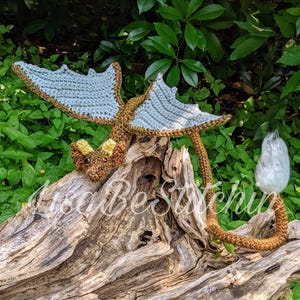 May include: A crocheted dragon figurine featuring a brown body, light blue wings, and yellow horns. The dragon has a long tail ending in a white tuft and is positioned on a piece of driftwood. The text LisaBeStitchin is visible across the image.