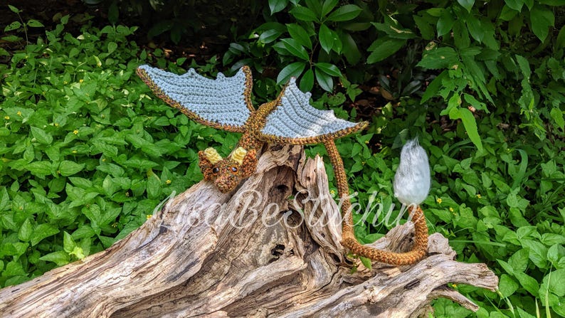 May include: A crocheted dragon figurine featuring a brown body, light blue wings, and yellow horns. The dragon has a long tail ending in a white tuft and is positioned on a piece of driftwood. The text LisaBeStitchin is visible across the image.