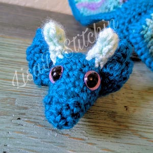 May include: A crocheted dragon toy featuring a deep blue body, light green ear-like accents, and large black eyes with pink irises. The creature has a textured yarn surface and a long tail extending backward. The text LisaStitchin is visible.