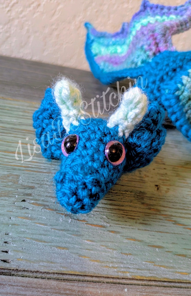 May include: A crocheted dragon toy featuring a deep blue body, light green ear-like accents, and large black eyes with pink irises. The creature has a textured yarn surface and a long tail extending backward. The text LisaStitchin is visible.